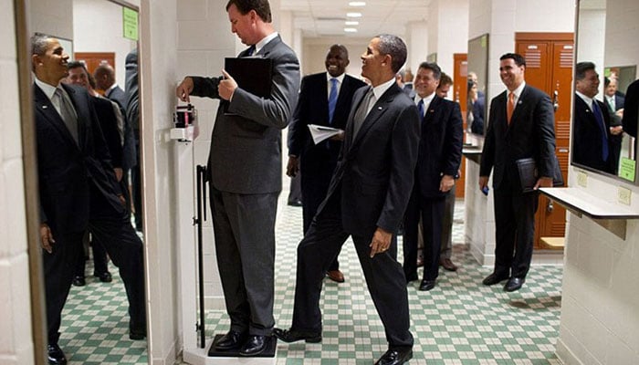 August 9, 2010. Obama puts his toe on the scale as Trip Director Marvin Nicholson tries to weigh himself during a hold in the volleyball locker room at the University of Texas