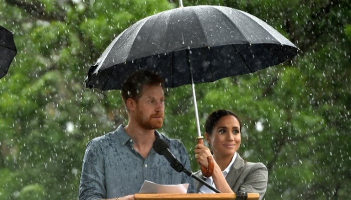 Meghan holds an umbrella as Prince Harry speaks to an outback community in a rare downpour