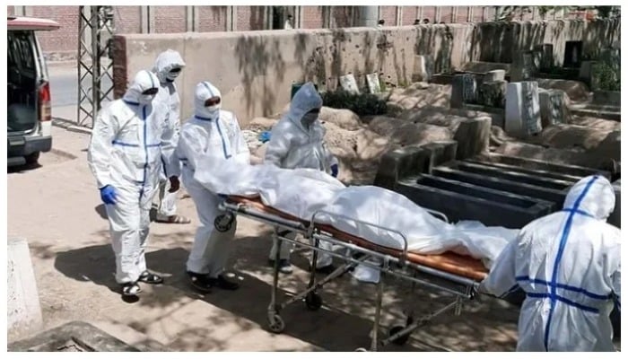 Paramedics dressed in Personal Protective Equipment carry a body on a stretcher inside a graveyard.