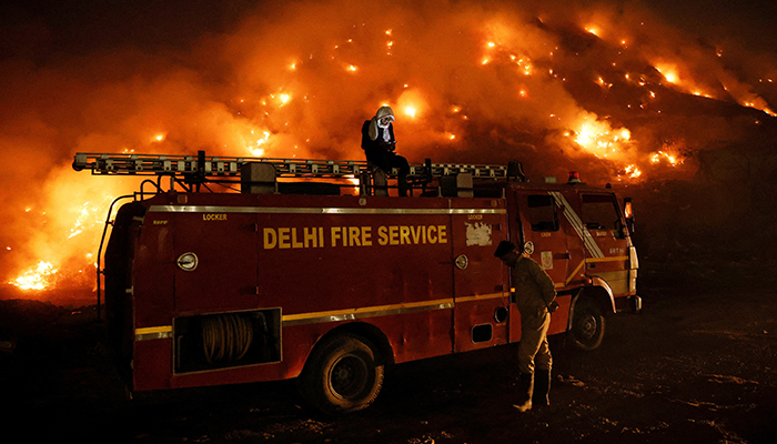 A firefighter uses his mobile phone as he sits on top of a fire truck as smoke billows from burning garbage at the Bhalswa landfill site in New Delhi, India, on April 27, 2022. — Reuters