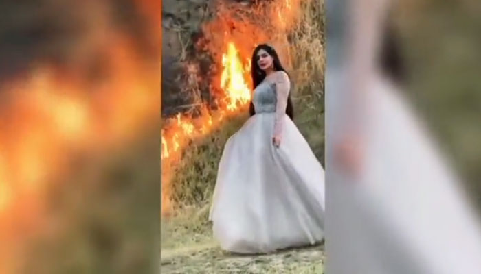 Woman posing for a photo as a fire rages in a field behind her. — Screengrab via Twitter/@rinasaeed