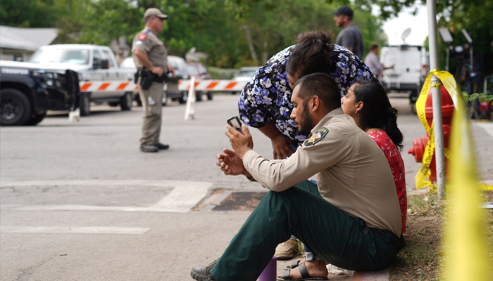 A sheriff checks his phone as he sits on the sidewalk with two women outside Robb Elementary School. Photo— allison dinner / AFP
