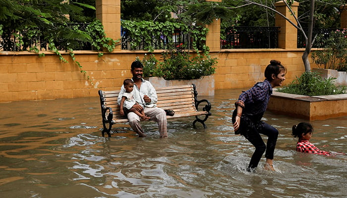Seorang pria dengan bayi duduk di bangku sementara anak-anak bermain di tengah jalan yang banjir selama musim hujan di Karachi, Pakistan, 11 Juli 2022. — Reuters