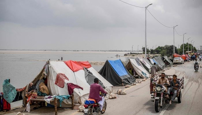 26 اگست، 2022 کو جنوبی پاکستان کے صوبہ سندھ کے سکھر میں مون سون کی شدید بارشوں کے بعد سیلاب زدہ گھروں سے بھاگنے والے بے گھر افراد کے لیے بنائے گئے عارضی خیموں کے سیٹ پر موٹرسائیکل سوار گزر رہے ہیں۔ — AFP