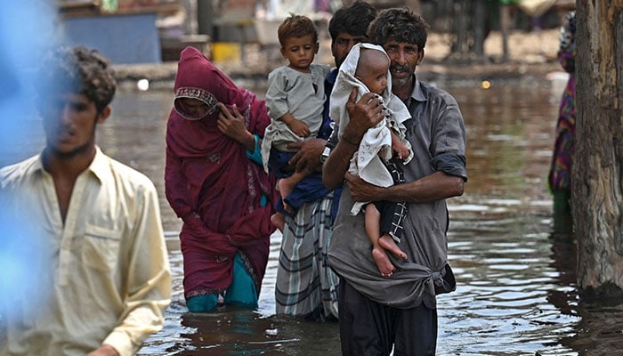7 ستمبر 2022 کو صوبہ سندھ کے ضلع دادو میں مون سون کی شدید بارشوں کے بعد ایک چھوٹے سے پاکستانی گاؤں بستی احمد دین میں پاکستانی بحریہ کی ایک کشتی پر بیٹھنے کے لیے پھنسے ہوئے سیلاب سے متاثرہ لوگ سیلابی پانی میں سے گزر رہے ہیں۔ — AFP