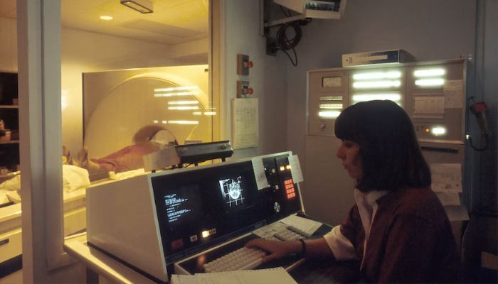 A computer-assisted tomographic (CAT) scanner, with a female technician working at a screen and behind a glass wall.— Unsplash