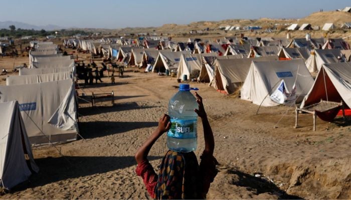 ایک بے گھر لڑکی پانی کی بوتل اٹھائے ہوئے ہے جو اس نے قریبی پھنسے ہوئے سیلابی پانیوں سے بھری تھی، جب اس کا خاندان سہون، پاکستان میں، 30 ستمبر 2022 کو ایک کیمپ میں پناہ لے رہا ہے۔— رائٹرز