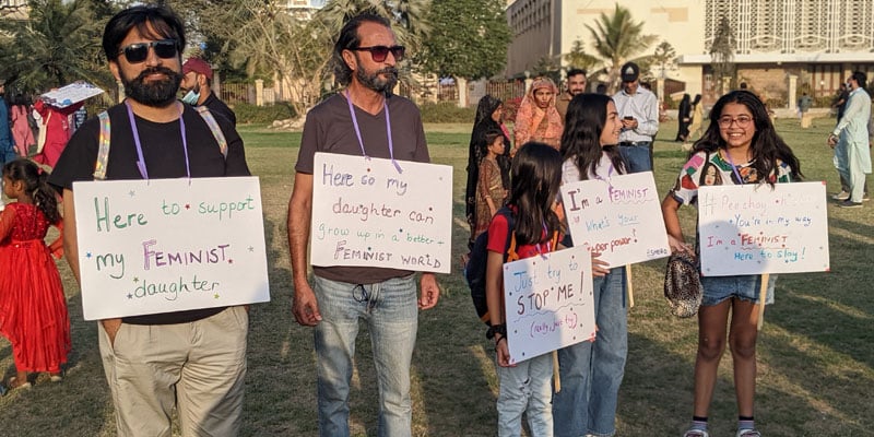 Two men with their daughters with placards hanged around their necks during Aurat March 2023 in Burns Garden Karachi on March 12, 2023. — Photo by author