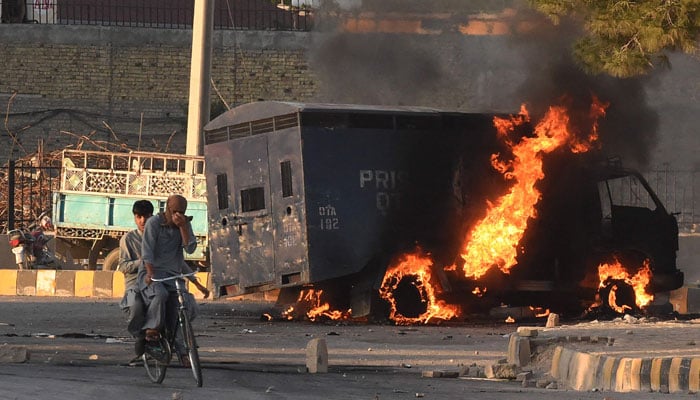 9 مئی 2023 کو کوئٹہ میں پی ٹی آئی کے کارکنوں اور سابق وزیر اعظم عمران خان کے حامیوں کی جانب سے اپنے رہنما کی گرفتاری کے خلاف احتجاج کے دوران موٹر سائیکل پر سوار مرد جلتی ہوئی پولیس گاڑی کے پاس سے گزر رہے ہیں۔ — اے ایف پی