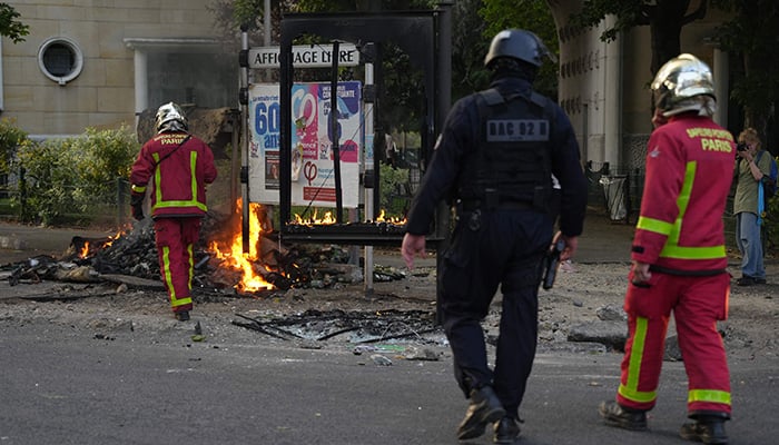 Firefighters walk with a police officer in riot gear as they arrive to put out fires after a demonstration in Nanterre, west of Paris, on June 27, 2023. — AFP