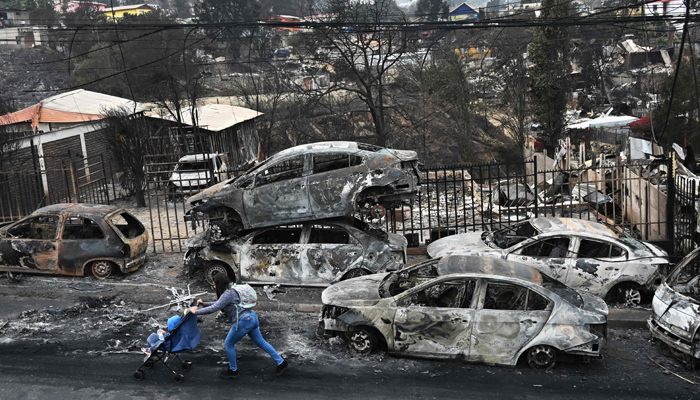 A woman with her baby walks past burned vehicles after a forest fire in Quilpue,Viña del Mar, Chile, on February 4, 2024. — AFP