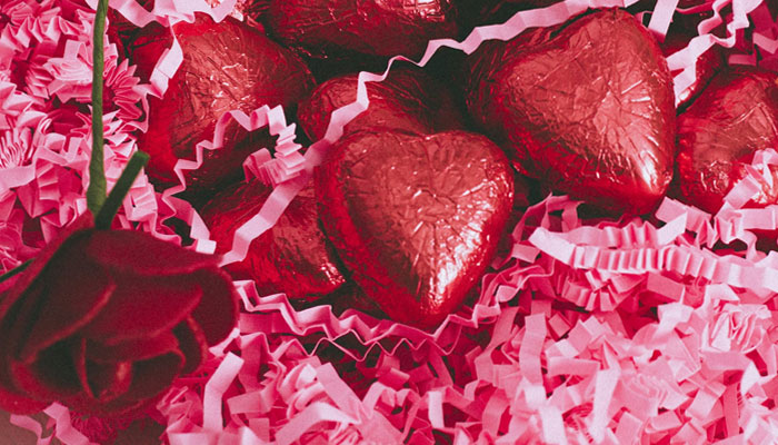 This image shows a bunch of wrapped heart-shaped chocolates on pink-coloured paper strips next to a fake rose. — Unsplash