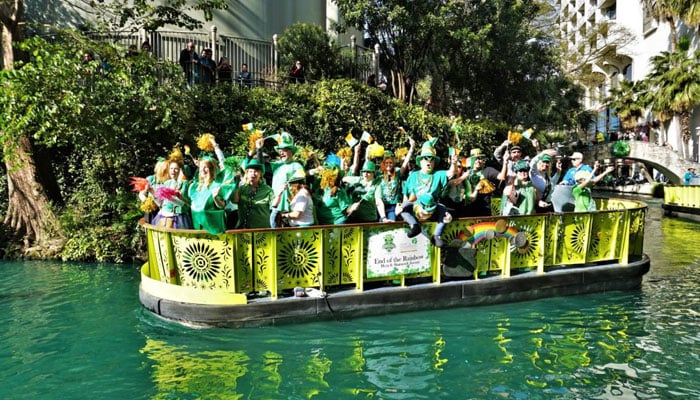 People dressed in St Patricks Day outfits join a parade on a float across the San Antonio River Walk. — Visit San Antonio