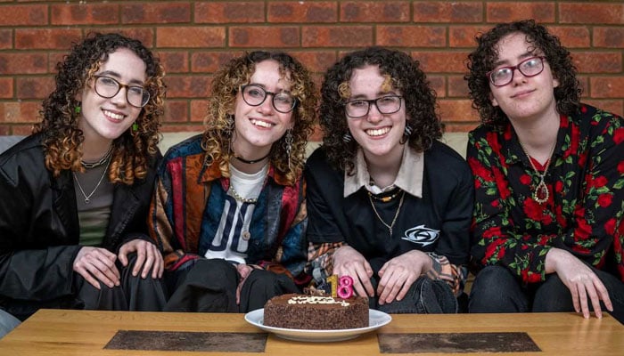 This image shows quadruplets Ellie, Holly, Georgie and Jess sitting together in front of a birthday cake. — Good News Network via SWNS