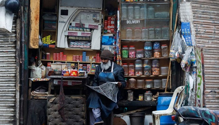 A man reads newspaper while selling betel leave, cigarettes and candies from a shop in Karachi. — Reuters/File
