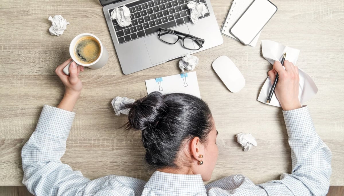 A representational image shows a woman resting her head on a desk due to stress. — Canva