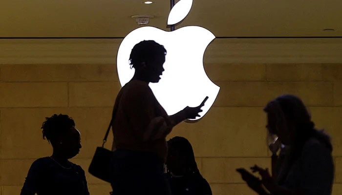 A woman uses an iPhone mobile device as she passes a lighted Apple logo at the Apple store at Grand Central Terminal in New York City, US, April 14, 2023. — Reuters