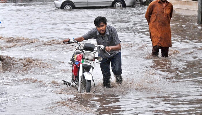 A young boy pushes his motorcycle through a rain-flooded road after a heavy downpour, highlighting the challenges faced by commuters in Multan, on June 26, 2025. — APP