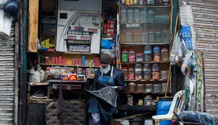 A man reads newspaper while selling betel leaves, known as pan, cigarettes and candies from a shop in Karachi. — Reuters/File