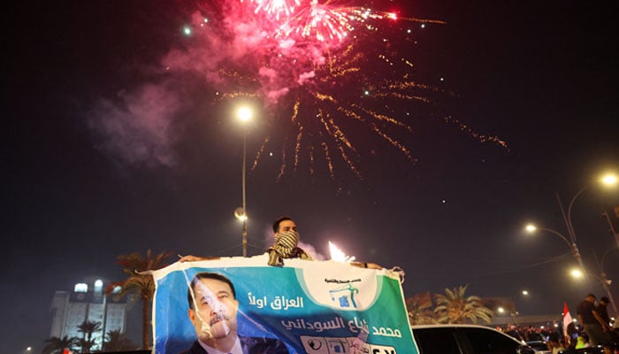 A man holds an election poster featuring current Prime Minister Mohammed Shia al-Sudani, with fireworks in the background, as supporters of the Reconstruction and Development Coalition celebrate after preliminary election results were announced in Baghdad, Iraq, November 12, 2025. — Reuters