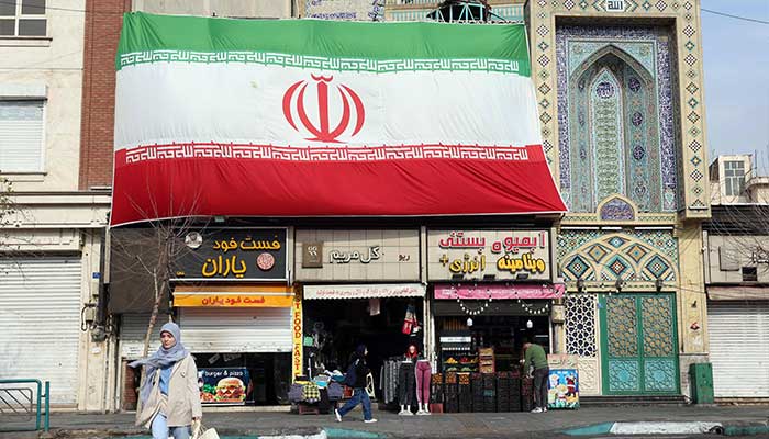 An Iranian woman walks past a huge national flag hanging above shops, in Tehran on February 6, 2026. — AFP