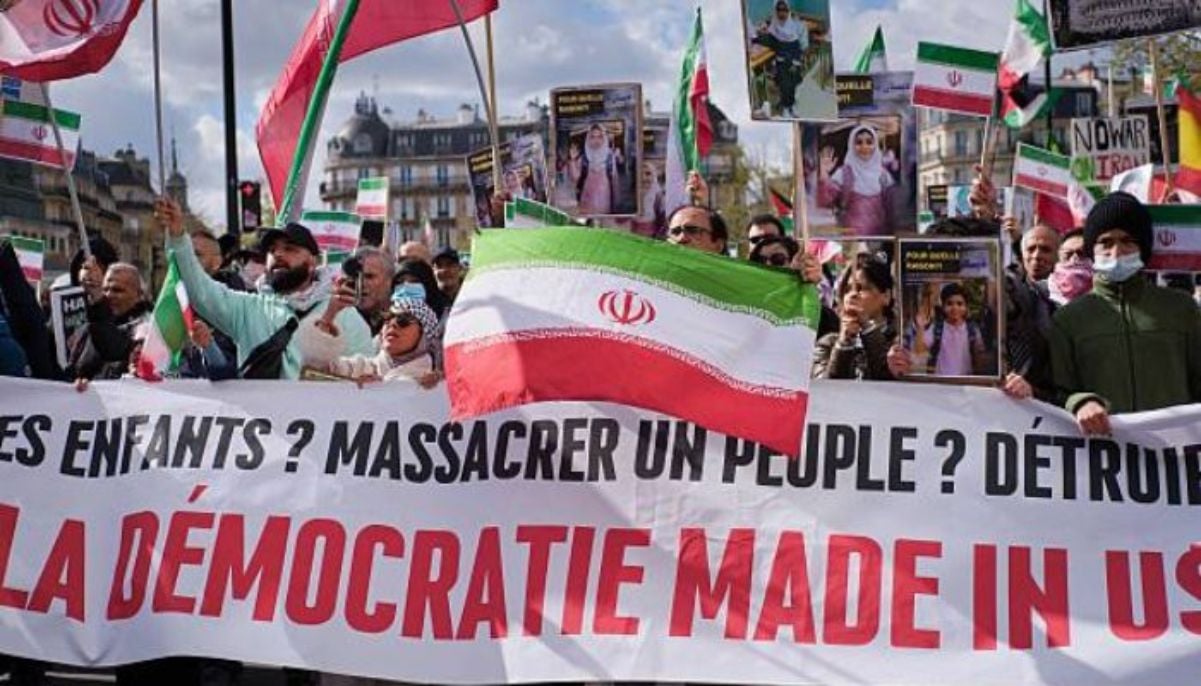 Wide-angle view of demonstrators holding banners and Iranian flags, including one with the large inscription DEMOCRACY MADE IN THE USA: MASSACRING A PEOPLE, DESTROYING A COUNTRY during a protest march in Paris, Ile-de-France, France, on March 28, 2026. — AFP
