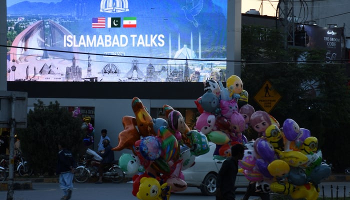 A balloon seller walks near a screen with an image referring to the U.S.-Iran peace talks on the day they are held in Islamabad, April 11, 2026.