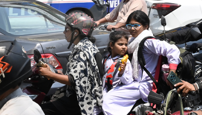 A woman rides a scooty with her children, while holding a mobile phone on a busy road after school in Lahore, on May 7, 2026. — APP