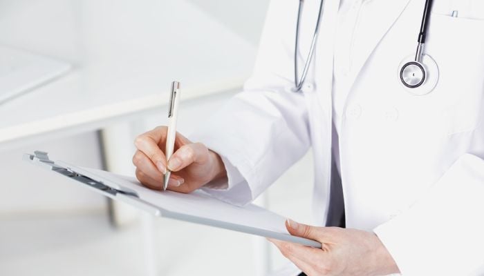 A doctor, with a stethoscope around his neck, writes on paper. —  Canva