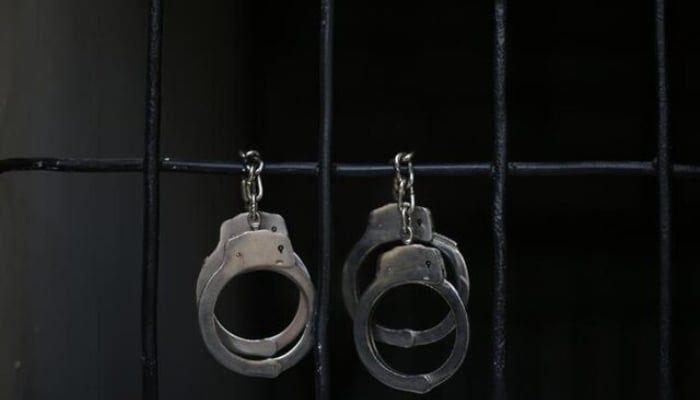 The image shows a pair of metallic steel handcuffs hanging from vertical dark prison or jail bars. — Reuters