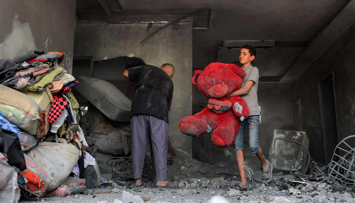 A boy carries a giant plush bear to a pile of salvaged items as people search the rubble of a collapsed building following overnight Israeli airstrikes in Gaza City on October 9, 2026. — AFP